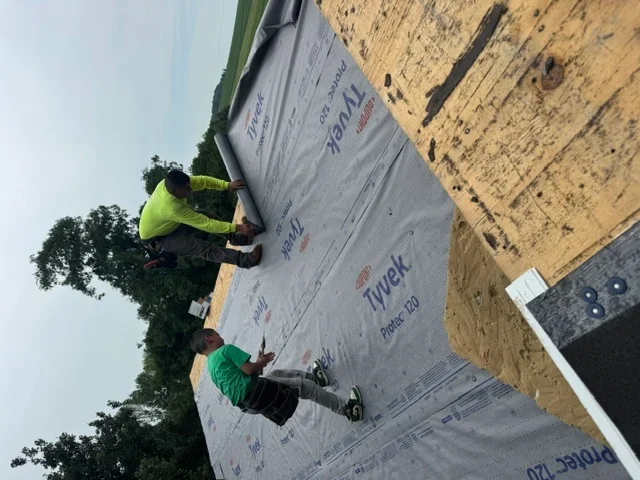 Crew installing gray synthetic underlayment on wooden roof deck with proper overlap